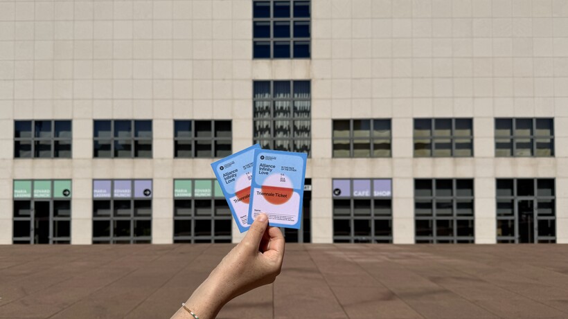 Hand hält zwei Tickets vor einem beigen Gebäude mit Fenstern